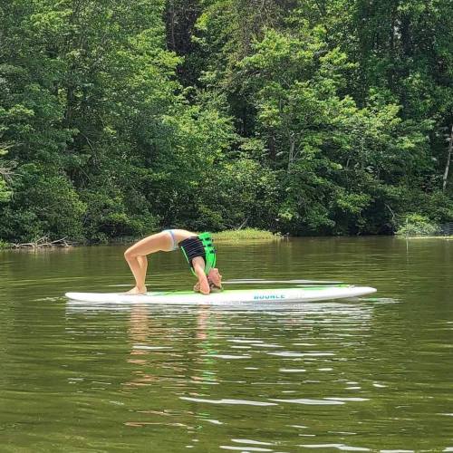 A person performs a yoga pose on a paddleboard in a calm, green lake surrounded by trees.