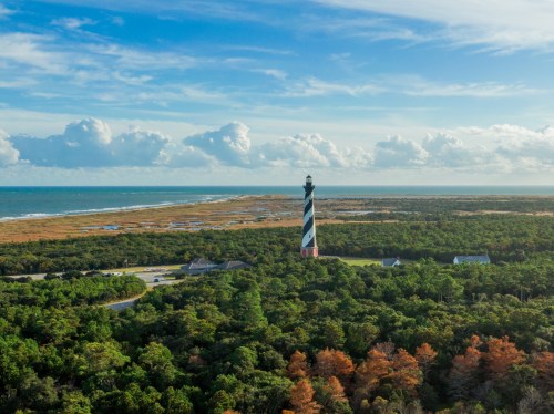 Aerial view of a striped lighthouse surrounded by greenery and coastline under a blue sky with fluffy clouds.