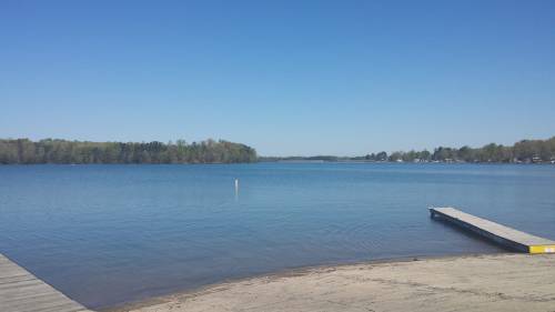 A calm lake under a clear blue sky, with a sandy shore and a wooden dock extending into the water.