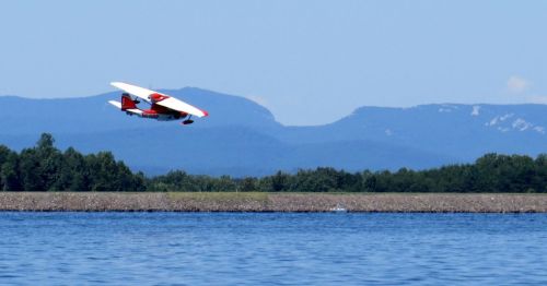 A seaplane flies low over a calm lake, with mountains in the background under a clear blue sky.