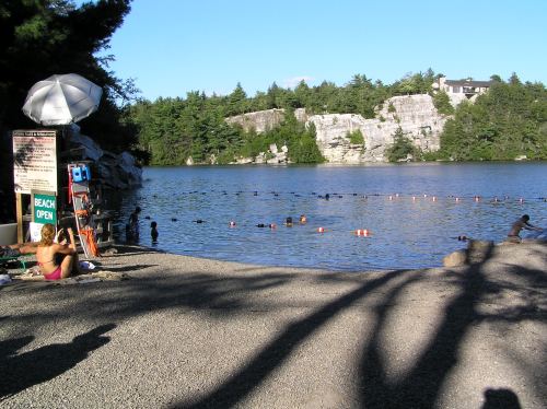 A sandy beach by a lake with swimmers, lifeguard equipment, and rocky cliffs in the background under a clear blue sky.