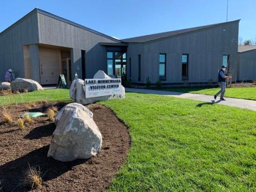 A modern visitor center with a stone sign, surrounded by green grass and landscaping, under a clear blue sky.