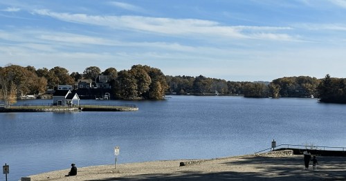 A serene lake scene with autumn trees, a small dock, and a person sitting on the shore under a clear blue sky.