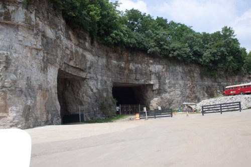 Two large rock tunnels cut into a cliffside, surrounded by greenery and a gravel area with a red bus in the foreground.