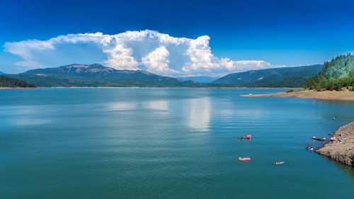 A serene lake surrounded by mountains, with fluffy clouds in a blue sky and people enjoying the water.