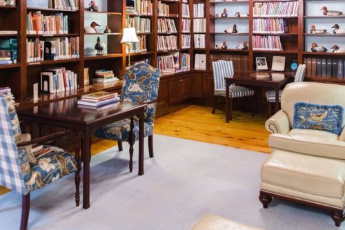 Cozy library with wooden shelves filled with books, two desks, and comfortable seating. Soft lighting enhances the atmosphere.