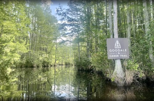 A serene view of a canoe trail surrounded by lush green trees and calm water, featuring a sign for Goodale State Park.