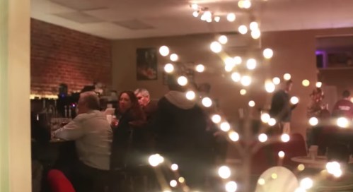 A cozy café scene with patrons enjoying drinks, illuminated by soft lights and a decorative tree in the foreground.