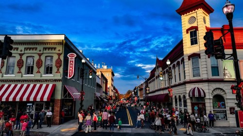 A vibrant street scene with shops and crowds under a blue sky, showcasing a lively atmosphere in a small town.