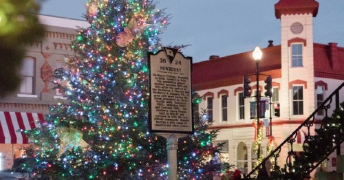 A brightly lit Christmas tree adorned with colorful lights, next to a historical marker in a festive downtown setting.