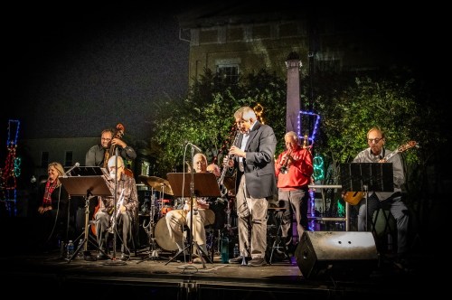 A jazz band performs on stage at night, surrounded by colorful lights and greenery. Musicians play various instruments.