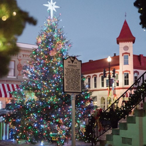 A brightly lit Christmas tree adorned with colorful lights stands in a festive town square at dusk.