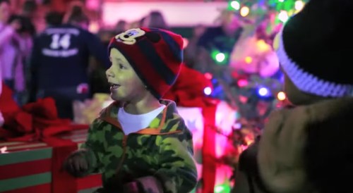A young child in a colorful hat smiles joyfully in front of a decorated Christmas tree and festive lights.