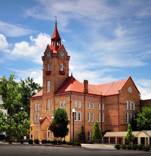 Historic brick building with a clock tower and red roof, surrounded by trees under a blue sky with clouds.
