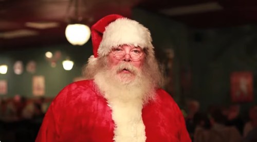 A man dressed as Santa Claus with a red suit, white beard, and glasses, standing in a festive setting.
