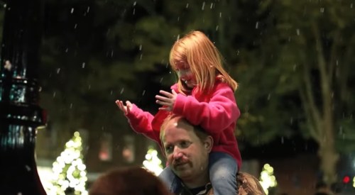 A child in a pink sweater sits on a man's shoulders, reaching out as snow falls around them.