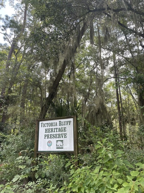 Sign for Victoria Bluff Heritage Preserve surrounded by lush greenery and hanging moss in a forested area.