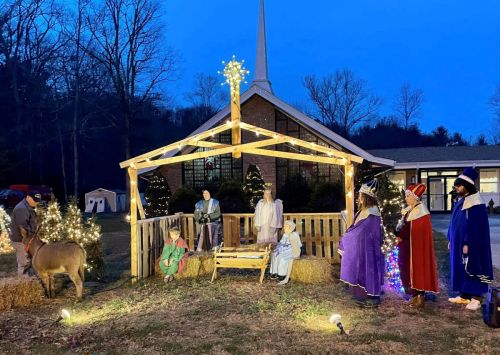 Nativity scene with figures, a donkey, and lights outside a church during twilight.
