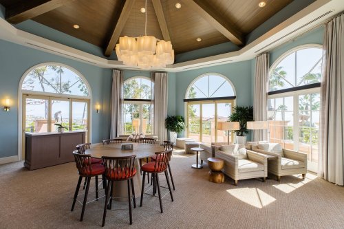 A spacious lounge with large windows, a round table, and comfortable seating, featuring a modern chandelier and coastal views.