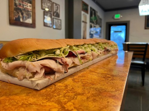 A long sub sandwich filled with layers of turkey, lettuce, and other toppings, resting on a table in a casual setting.