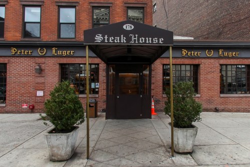 Entrance of Peter Luger Steak House, featuring a black awning and brick facade, with potted plants on either side.