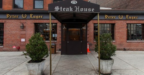 Entrance of Peter Luger Steak House, featuring a canopy and potted plants, with a brick facade and large windows.