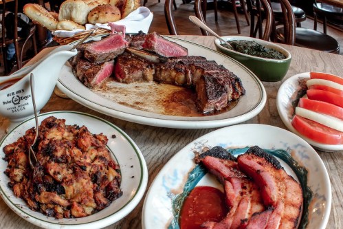 A table set with a platter of steak, sides of stuffing, greens, bacon, and sliced tomatoes, with bread rolls in the background.