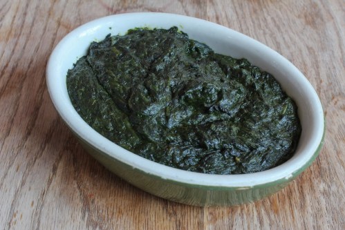 A green, thick dish of spinach or leafy greens served in an oval ceramic bowl on a wooden surface.