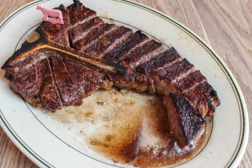 A perfectly cooked T-bone steak on a white plate, garnished with a pink cow-shaped pick, resting in its juices.