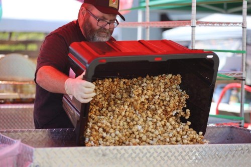 A person in a cap pours freshly made popcorn from a large container at a food stall.