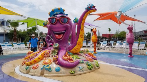 Colorful octopus fountain with sunglasses, surrounded by playful water features and lounge chairs at a pool area.
