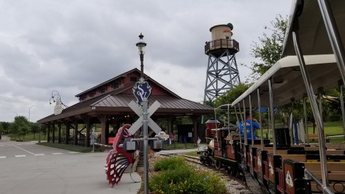 A train station with a wooden building, a water tower, and a train ready to depart, set against a cloudy sky.