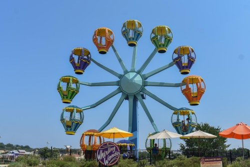 A colorful Ferris wheel with yellow and green gondolas against a clear blue sky, surrounded by umbrellas and amusement park scenery.