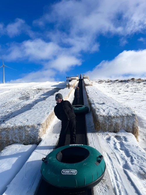 A person in winter gear stands on a snowy slope, holding a tube, with a blue sky and wind turbine in the background.