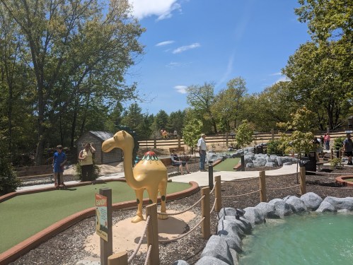 A mini-golf course featuring a golden camel statue, surrounded by trees and players enjoying the game on a sunny day.