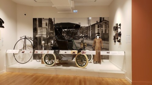 A vintage car displayed in a museum, alongside a bicycle and historical photographs on the wall.