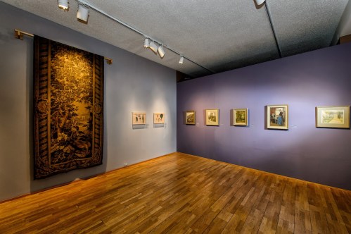 Art gallery interior featuring framed artworks on a purple wall and a large tapestry hanging on the left. Wooden floor.