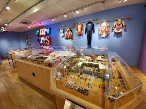 A colorful exhibit featuring a miniature circus scene and vintage costumes under bright lights in a gallery.