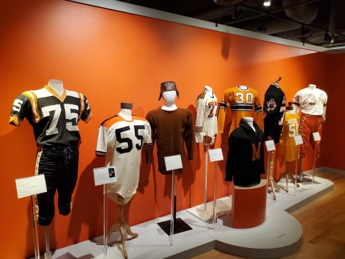 A display of vintage sports uniforms on mannequins against an orange wall, showcasing various styles and numbers.