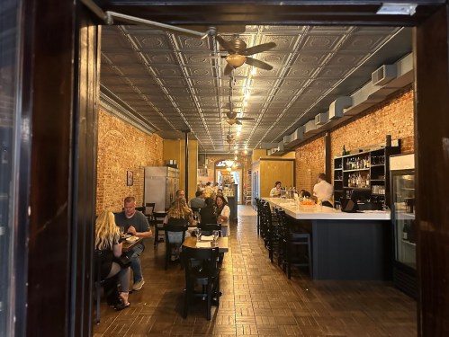 View of a restaurant interior with exposed brick walls, a bar, and patrons dining at tables.