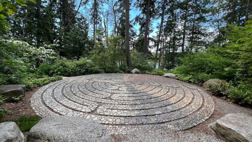 A stone labyrinth surrounded by lush greenery and trees, set in a tranquil outdoor environment.