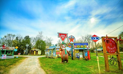 A whimsical roadside scene featuring vintage gas station signs and a large cartoon bear statue, set against a blue sky.