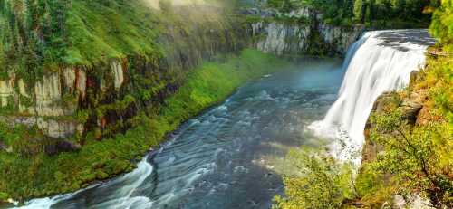 A stunning waterfall cascades over rocky cliffs, surrounded by lush greenery and mist rising from the flowing river below.