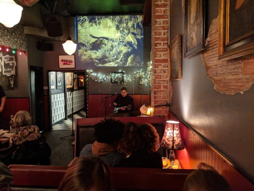 A cozy venue with a musician performing on stage, surrounded by dim lighting and a nature-themed projection on the wall.