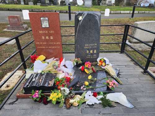 Two gravestones for Bruce Lee and Brandon Lee, surrounded by flowers and offerings in a serene cemetery setting.