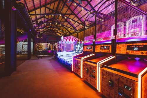 A vibrant arcade with neon lights, featuring games like skee-ball and a carnival booth in a spacious, rustic setting.