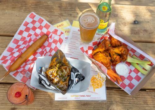 A table with a variety of food: nachos, chicken wings, a corn dog, a drink, and a cocktail, all on checkered paper.