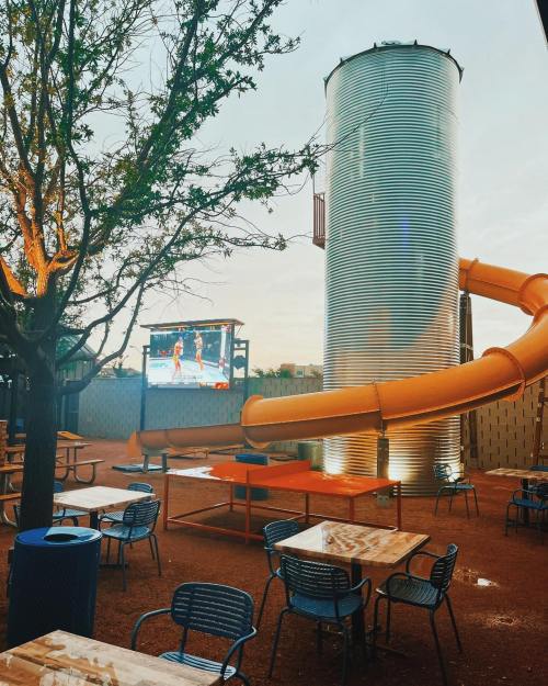 Outdoor seating area with tables, a large screen for viewing, a metal silo, and a bright orange slide.
