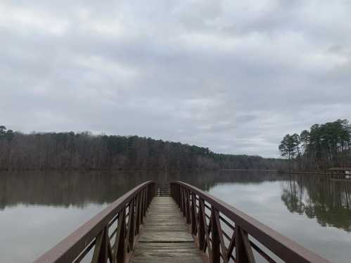 A wooden bridge extends over a calm lake, surrounded by trees under a cloudy sky.