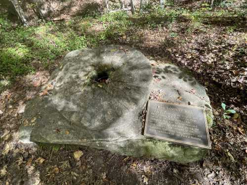 A large stone millstone sits on the ground, accompanied by an informational plaque in a wooded area.